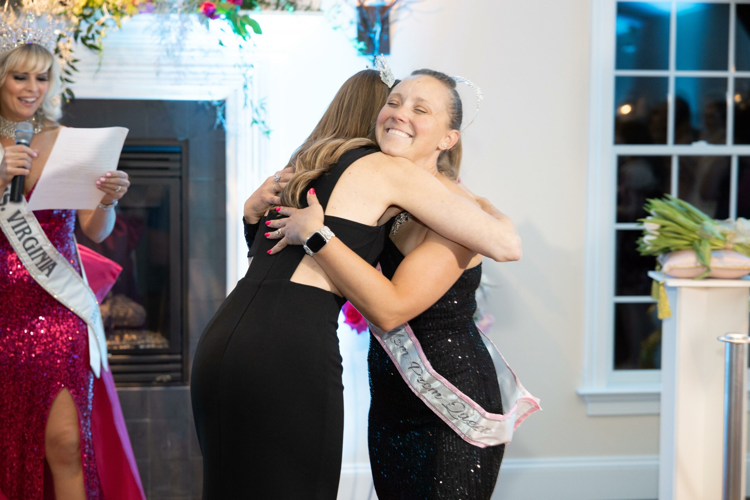 A young woman with a crown and sash stands smiling with her arms wrapped around another woman who is wearing a queen sash and crown.